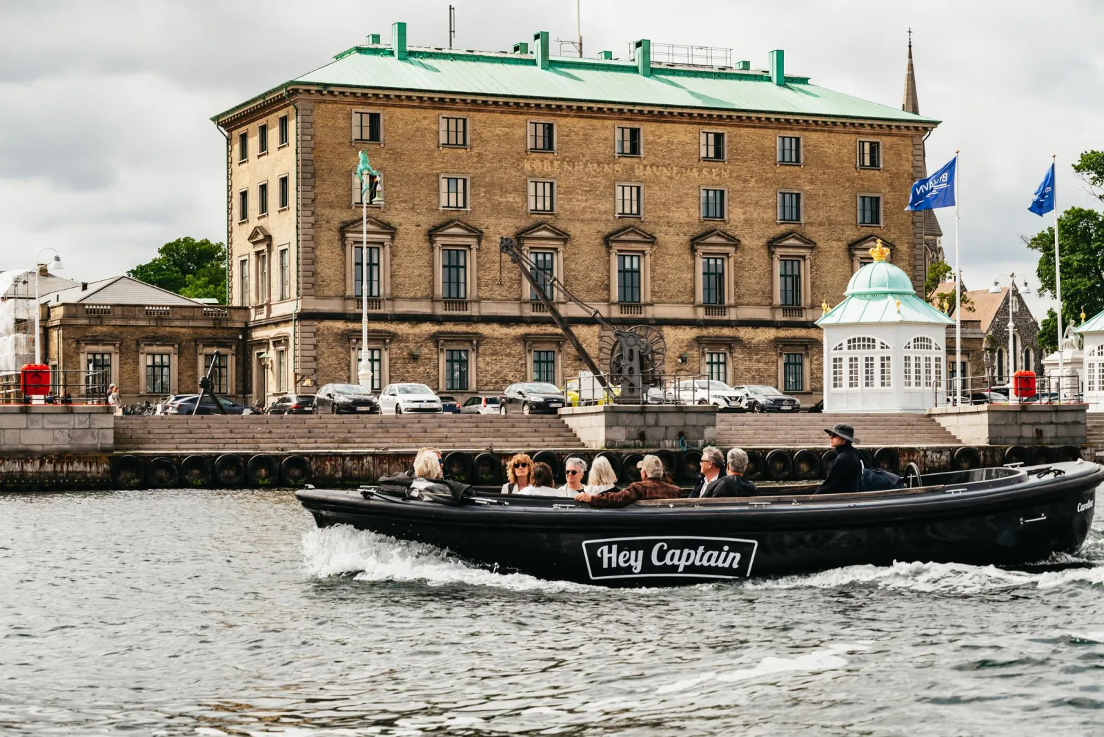 A Hey Captain boat passing a historic red-brick building with a green roof