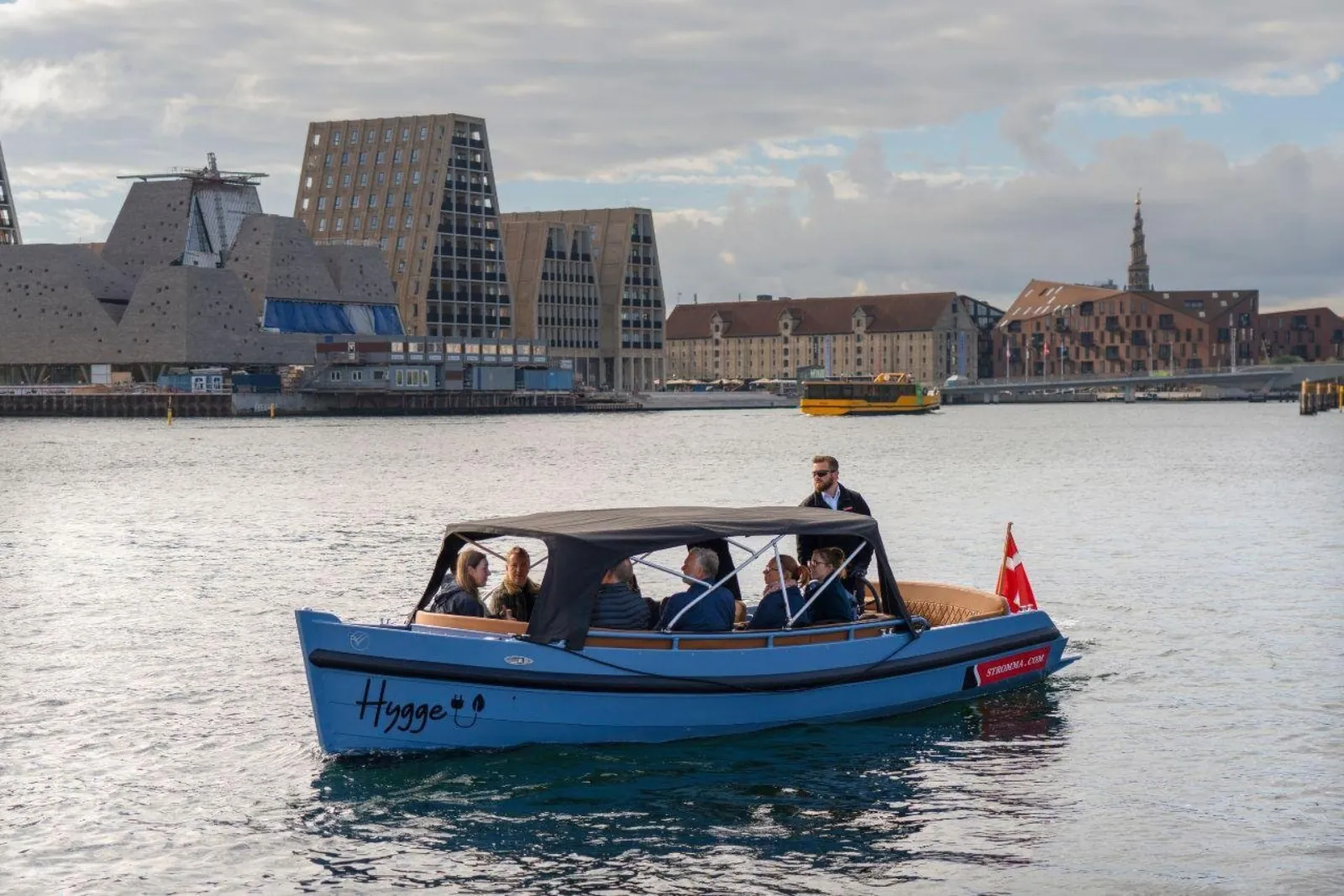 A blue electric boat passing modern harbourfront buildings