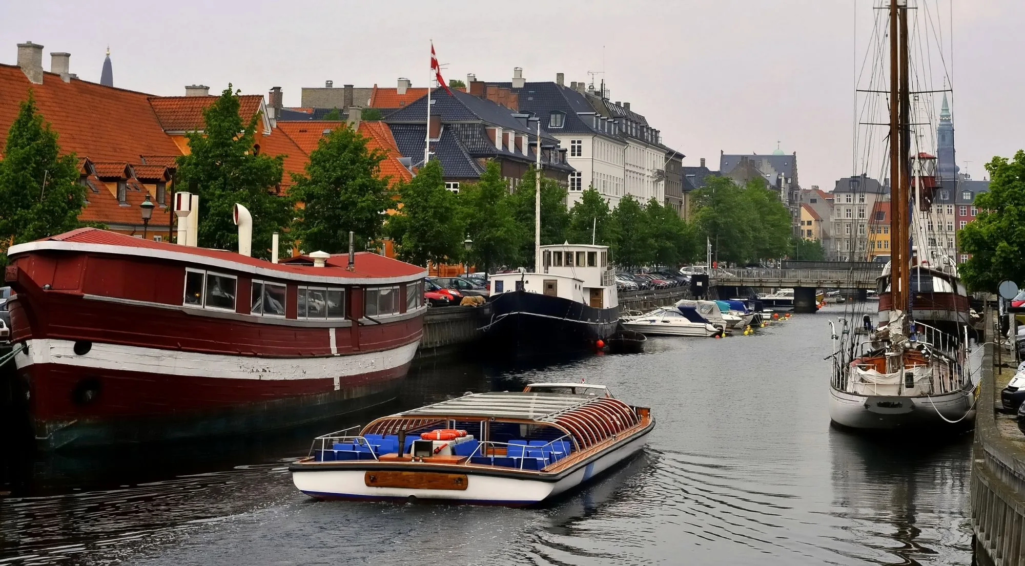 An open canal boat passing old wooden ships in a Copenhagen canal